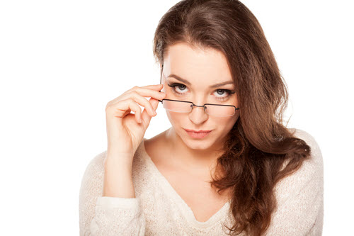 A woman with long brown hair and a light sweater looks directly at the camera, slightly raising her black-rimmed glasses with one hand. She has a confident, inquisitive expression. The background is white and uncluttered.