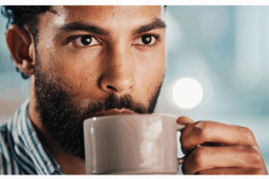 A close-up of a bearded man wearing a striped shirt, holding a white mug with both hands and sipping from it. His expression is thoughtful, and bright natural light softly blurs the background. A close-up of a bearded man wearing a striped shirt, holding a white mug with both hands and sipping from it. His expression is thoughtful, and bright natural light softly blurs the background.