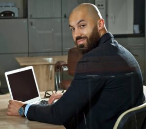 A bearded man with a shaved head, wearing a dark jacket and blue watch, sits at a desk with an open laptop. He looks over his shoulder and smiles at the camera. The background shows office furniture and a glass cabinet. A bearded man with a shaved head, wearing a dark jacket and blue watch, sits at a desk with an open laptop. He looks over his shoulder and smiles at the camera. The background shows office furniture and a glass cabinet.