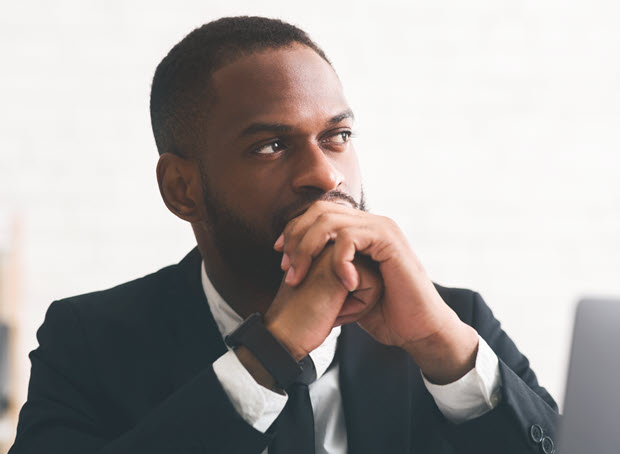 A man in a suit sits at a desk, hands clasped in front of his face, looking thoughtfully to the side. He has short hair, a trimmed beard, and wears a smartwatch. The background is softly blurred, creating a focused, contemplative atmosphere.