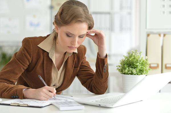 A woman wearing a brown blazer sits at a desk, writing in a notebook with a pen. She appears focused and thoughtful, with her head resting on one hand. An open laptop and a small potted plant are on the white desk in front of her.