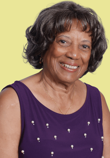 Brenda Goburn Smith, an older woman with short, curly gray hair, smiles warmly. She is wearing a sleeveless purple top with pearl-like embellishments and a delicate gold necklace. The background is a solid light yellow. Brenda Goburn Smith, an older woman with short, curly gray hair, smiles warmly. She is wearing a sleeveless purple top with pearl-like embellishments and a delicate gold necklace. The background is a solid light yellow.